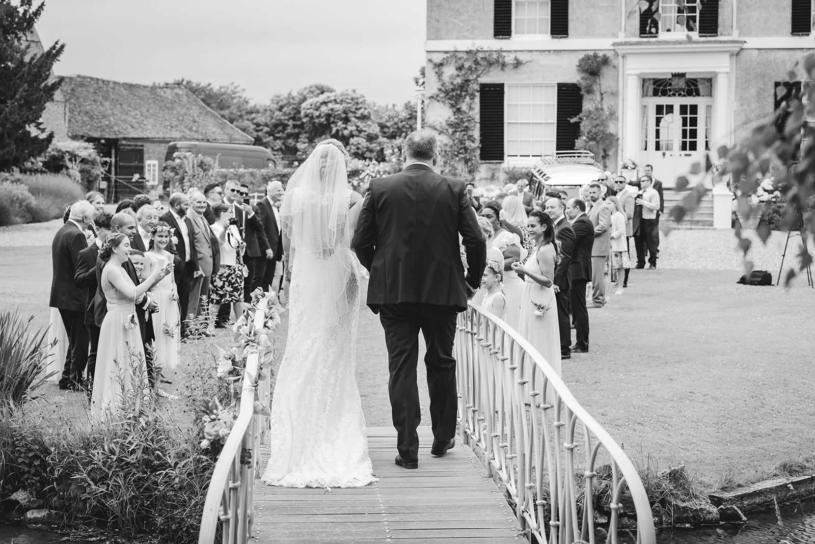 Newlyweds walk away across a decorative bridge towards a large country house while wedding guests look on from the lawn.
