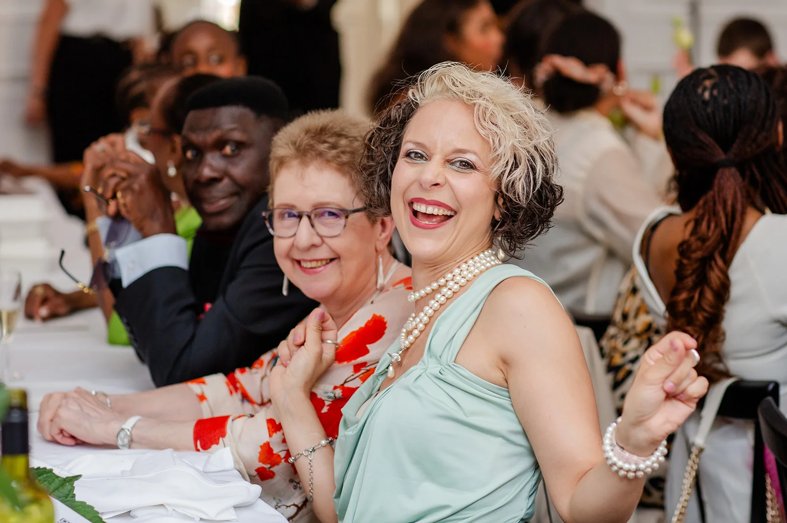 A joyful wedding guest with styled silver and dark hair laughs enthusiastically while seated at a reception table wearing a pale top and layered pearl necklaces.