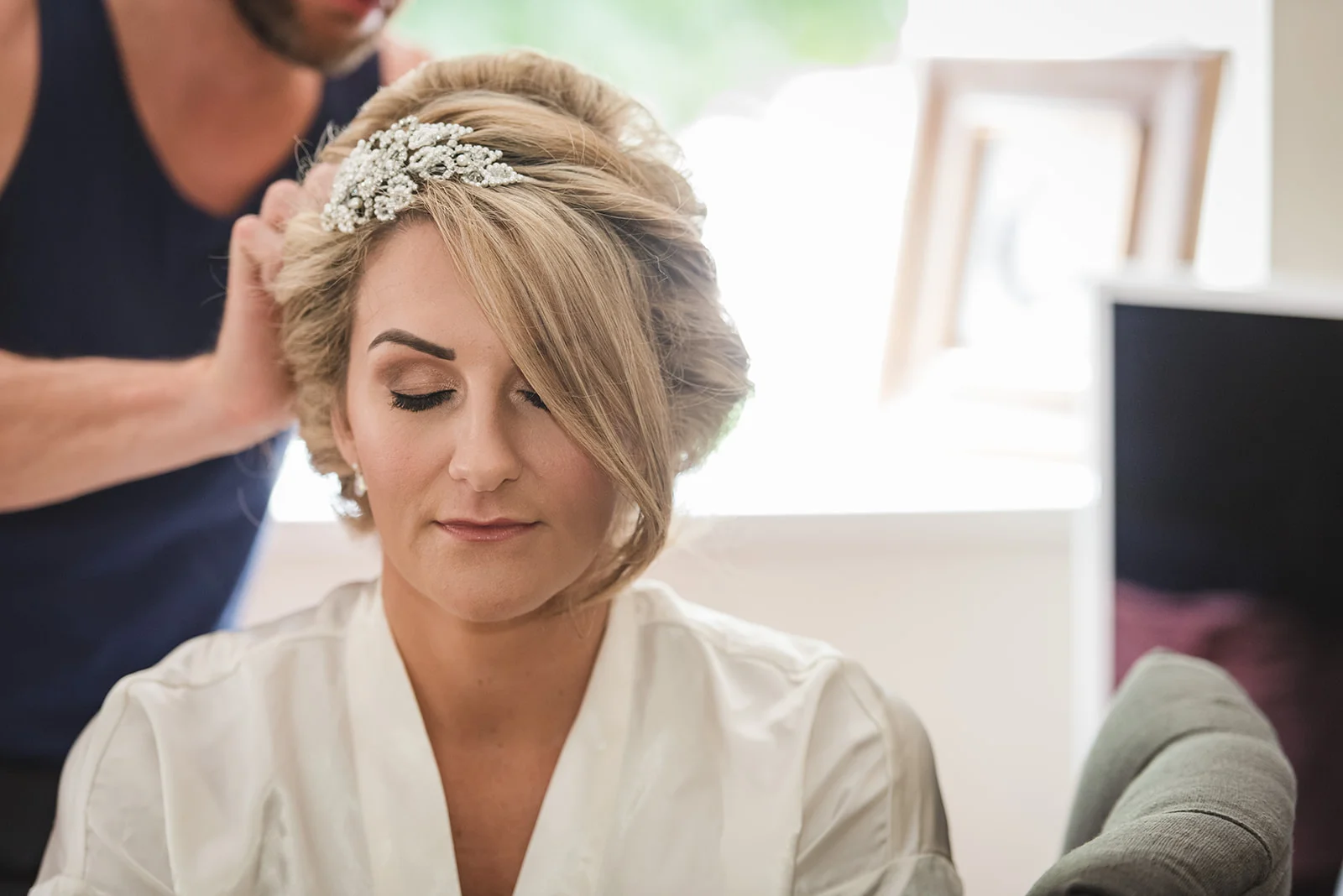 A bridal stylist carefully secures a sparkling crystal and pearl hair comb into the blonde hair of a bride during morning preparations.