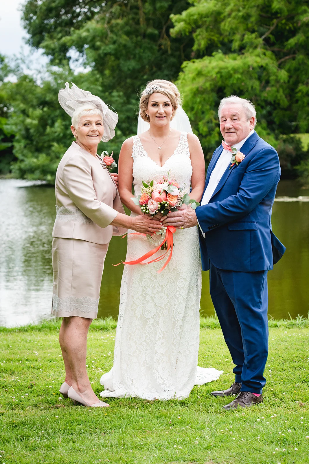 A smiling bride stands proudly outdoors by a lake, flanked by her mother in a chic skirt suit and her father in a blue suit, all holding her peach and pink bridal bouquet.