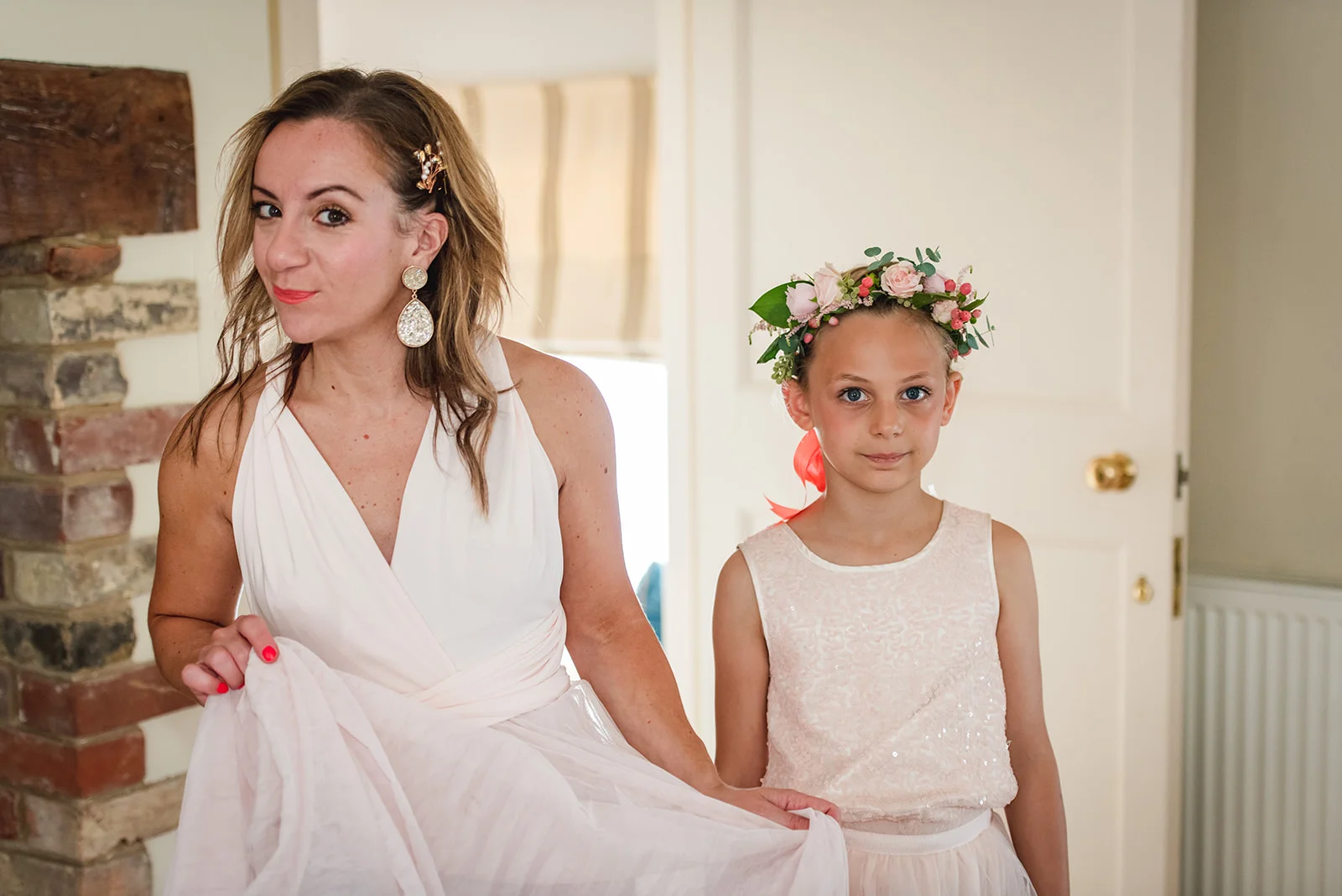 A woman wearing a pale pink dress and statement earrings stands beside a young flower girl wearing a floral crown during morning wedding preparations.