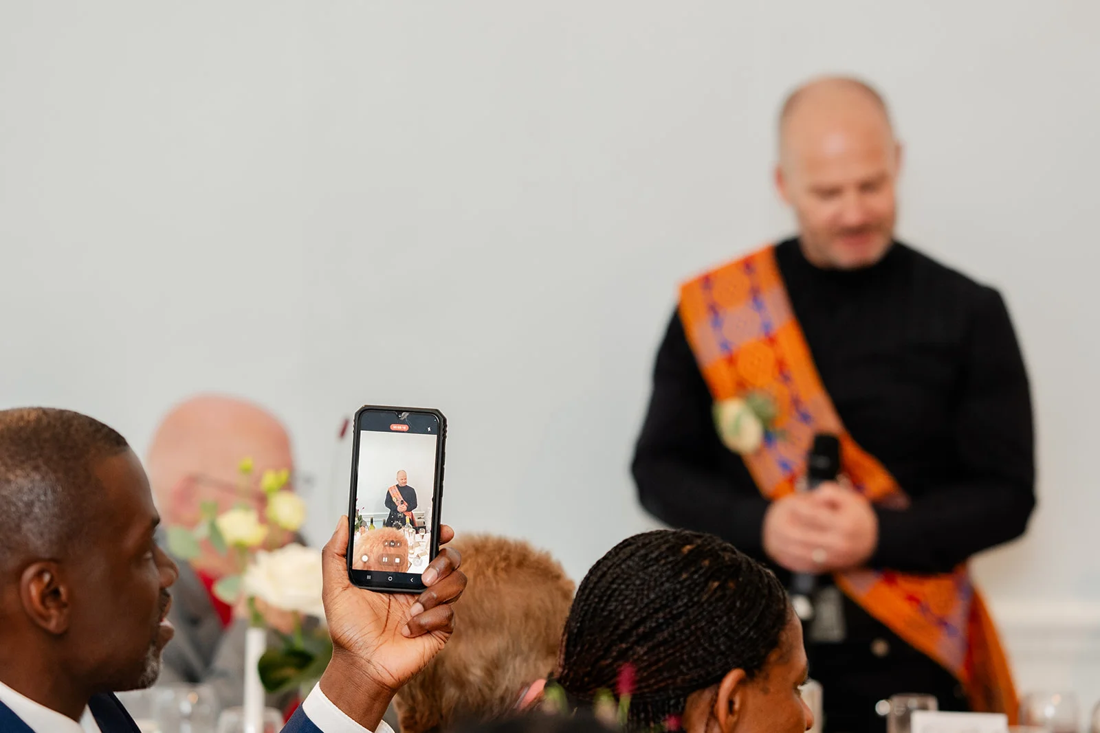 A wedding guest holds up a mobile phone to capture a speech being given by a person wearing a traditional tartan sash.