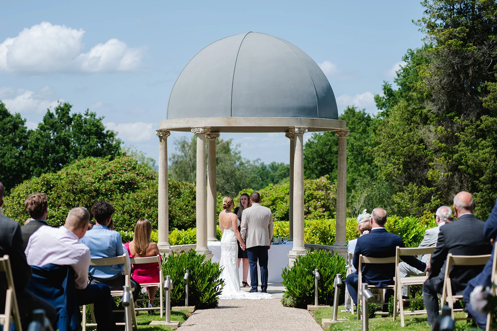 A bride and groom stand under a stone gazebo during an outdoor wedding ceremony while guests watch from seated rows on a sunny day.