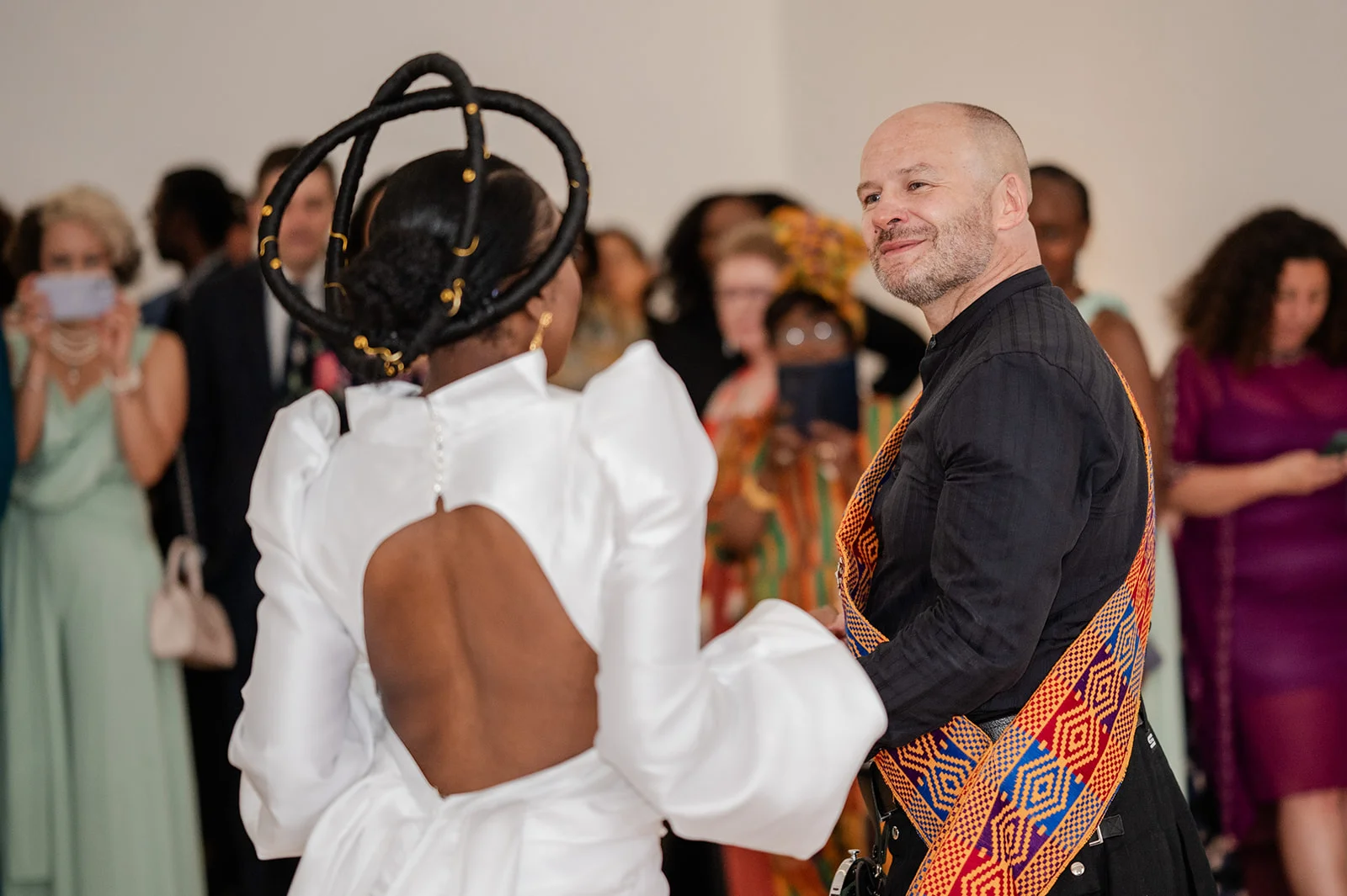An interracial couple smiling at each other during a wedding celebration, with the bride wearing a striking white gown featuring a keyhole back and a sculptural ring headpiece.