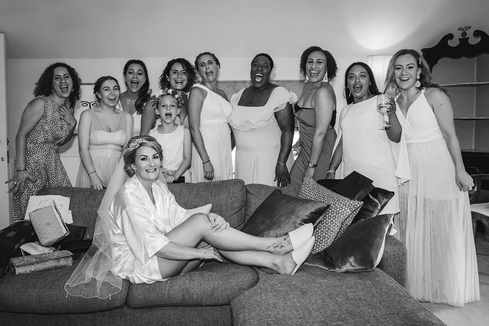 A bride in a white satin robe and veil sits smiling on a sofa, surrounded by a laughing group of bridesmaids and a flower girl cheering during morning preparations.