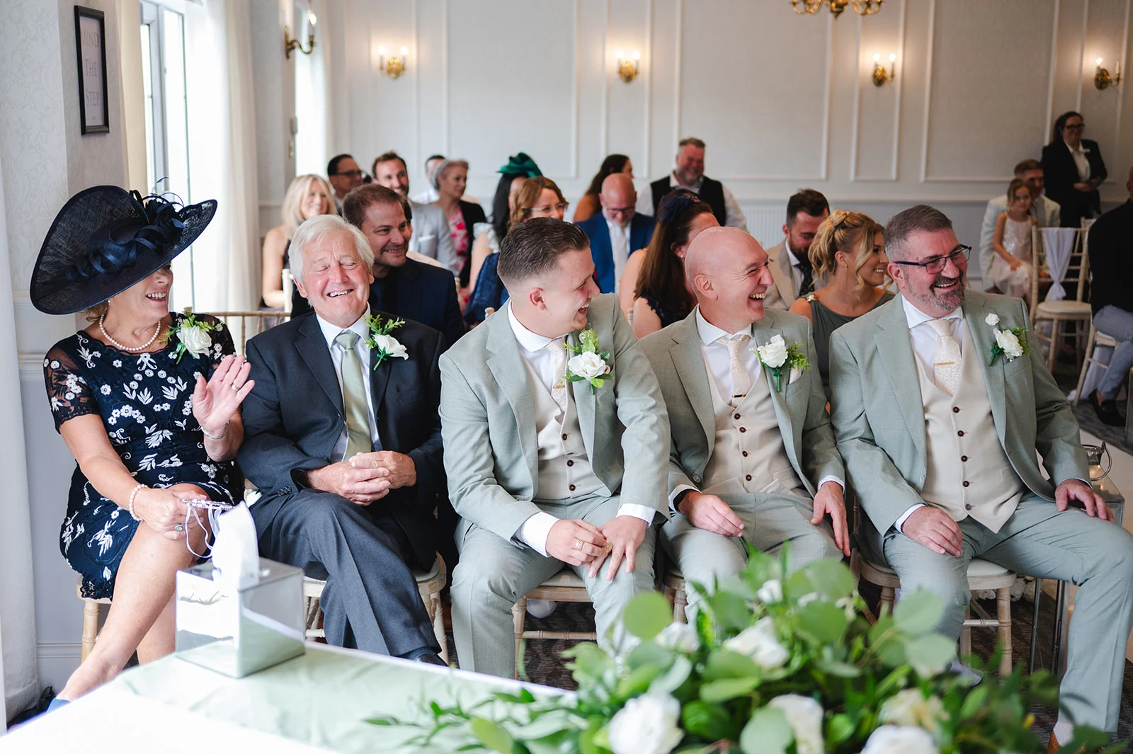 A joyful wedding party and guests seated at the front of a light-filled ceremony room, laughing heartily and sharing a humorous moment during the proceedings.