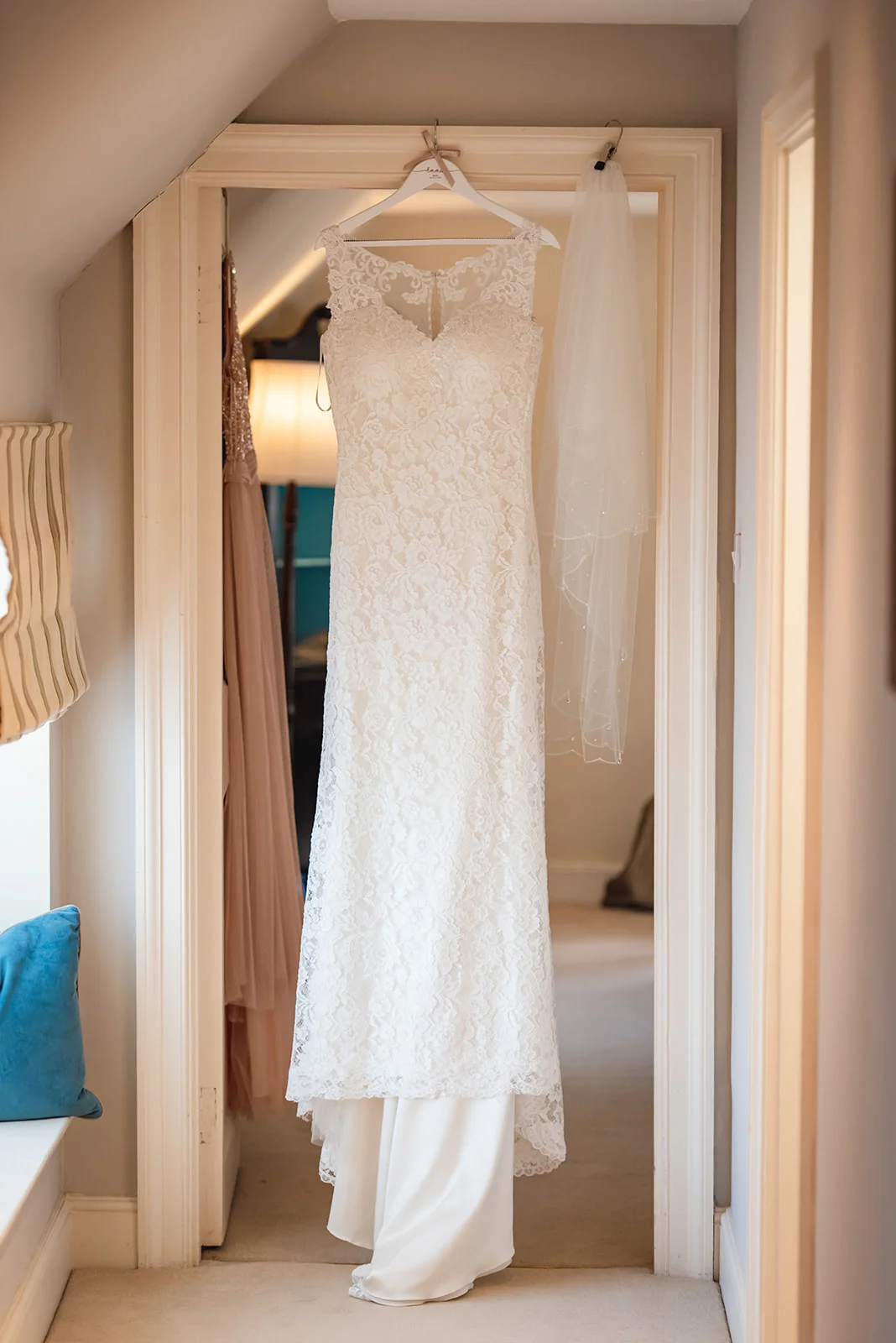 An intricate lace wedding dress with an illusion neckline hangs neatly in a bright doorway alongside a delicate veil during morning preparations.