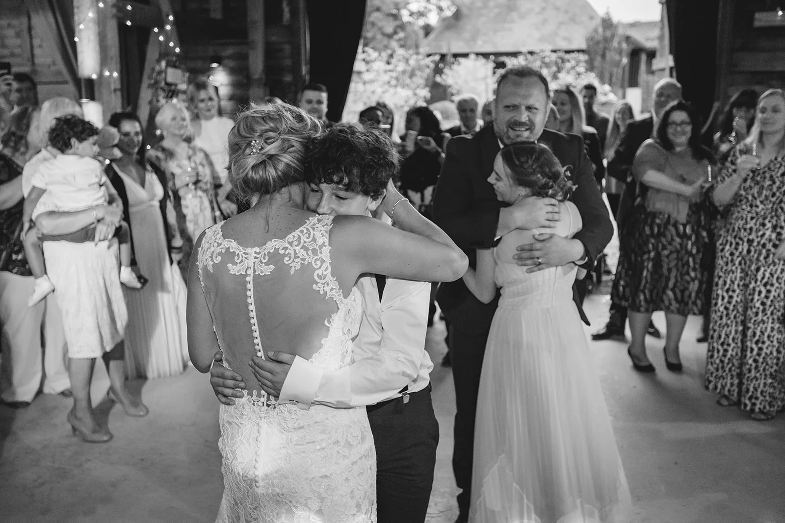A bride in a lace gown with a buttoned illusion back shares a tender embrace during a barn wedding celebration surrounded by family and guests.