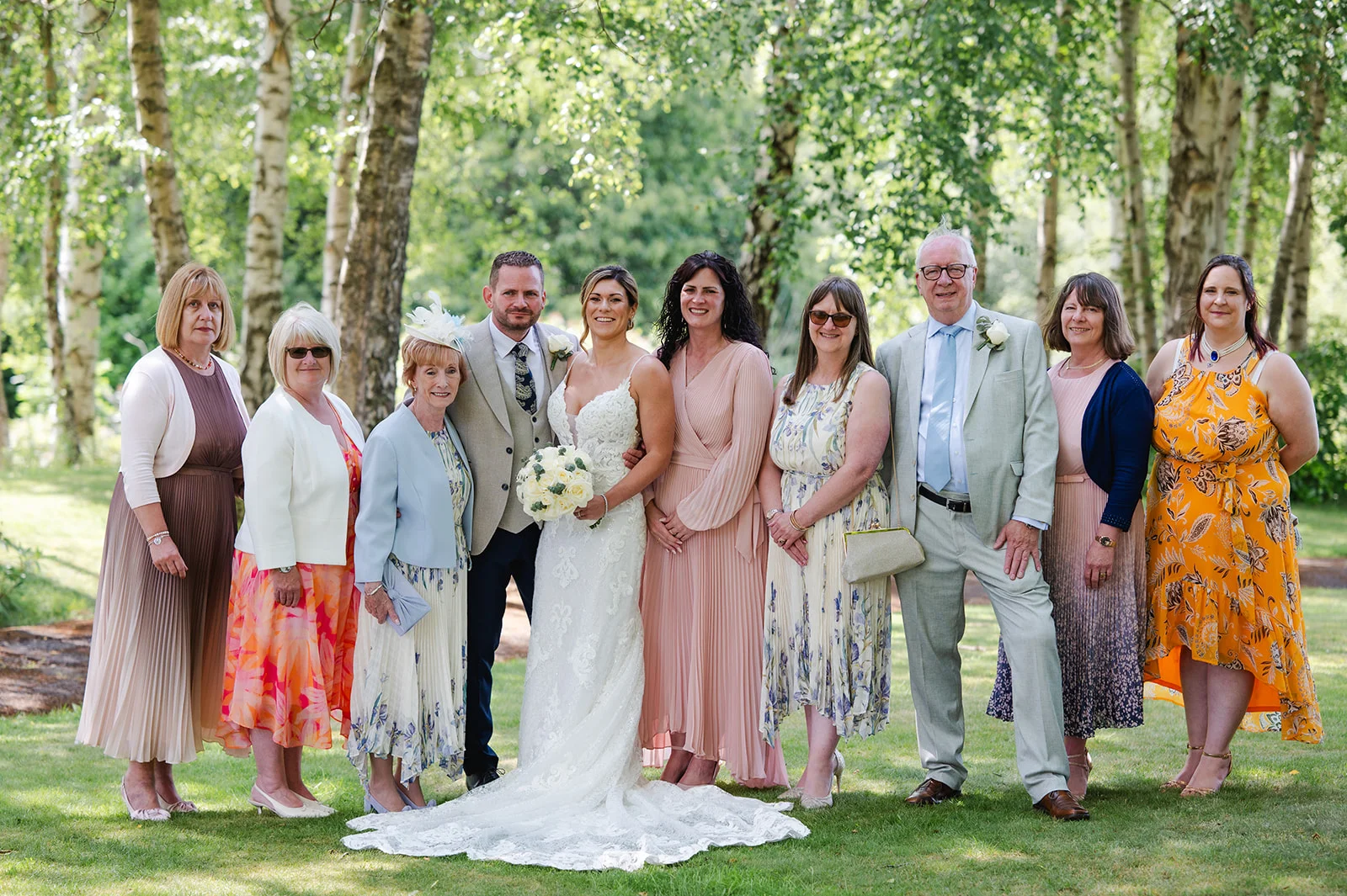 A wide shot shows the bride and groom posing outdoors in a sunlit woodland setting alongside a large group of family and wedding guests.