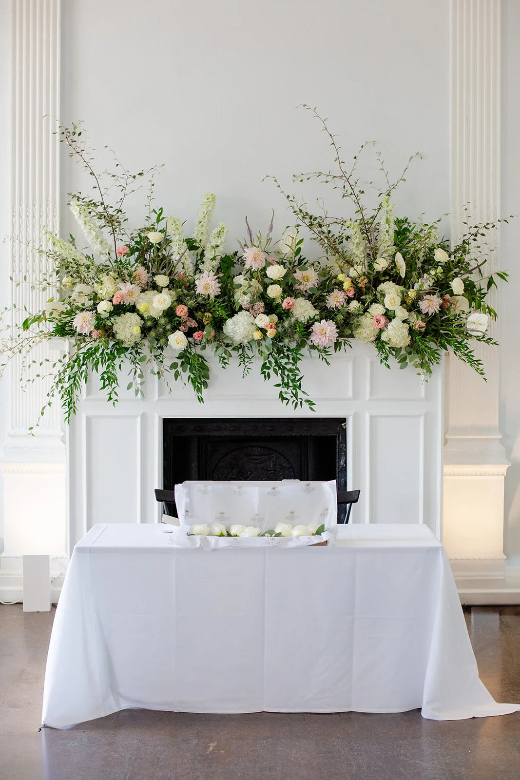 A white fireplace mantel adorned with an abundant floral installation of white and soft pink blooms, paired with a neatly dressed ceremony table below.