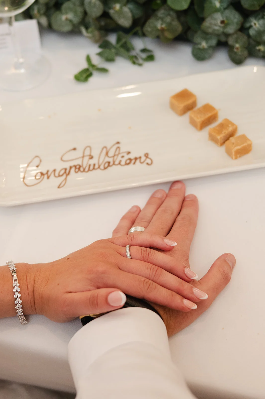 Close-up of the newlyweds holding hands on a white table next to a decorative plate with the word congratulations written on it.