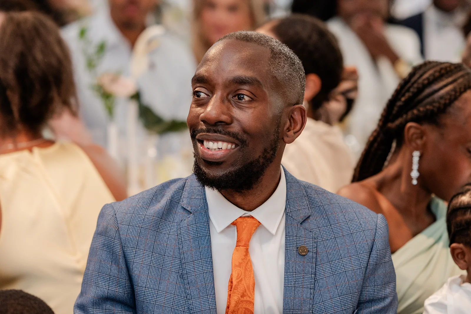 A smiling wedding guest in a checked blue suit and orange tie laughs joyfully during the reception.