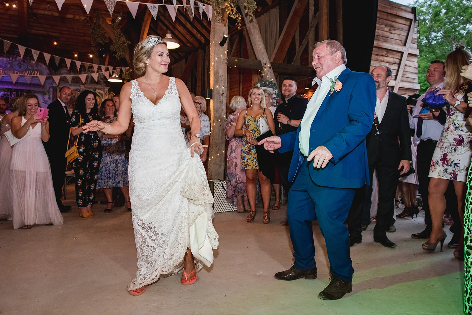A bride in a lace wedding dress and bright orange trainers shares a joyful dance with her father inside a rustic stone barn adorned with bunting and fairy lights.