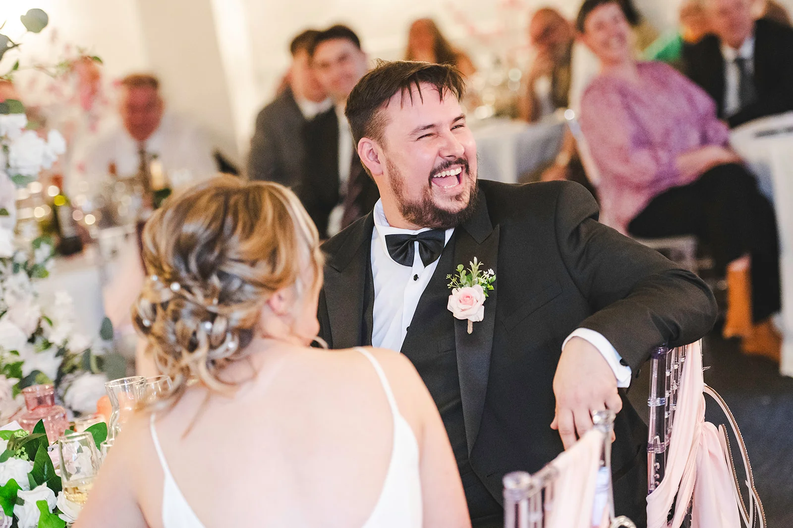 A groom in a classic black tuxedo laughs heartily during wedding speeches while sitting beside the bride at the top table.