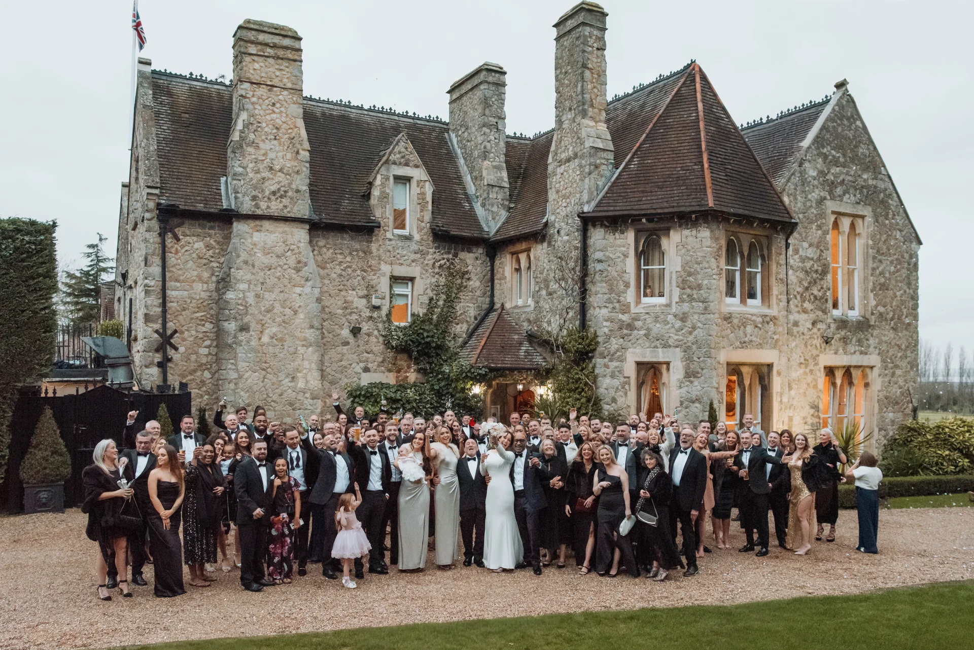 A large wedding party and guests gather for a celebratory toast outside the stone facade of a historic country house venue.