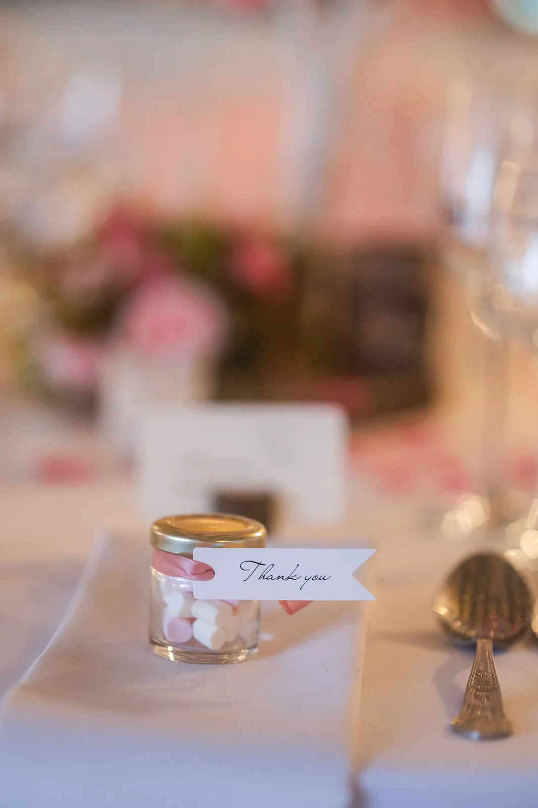 A sweet wedding favour jar filled with pink and white treats sits neatly on a crisp white napkin alongside polished cutlery.