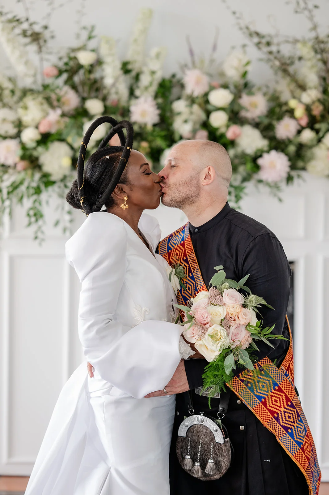 A newlywed interracial couple share a tender kiss in front of a large floral installation, with the bride wearing a striking white gown with balloon sleeves and a sculptural headpiece, and the groom wearing a black kilt with a colourful patterned sash.