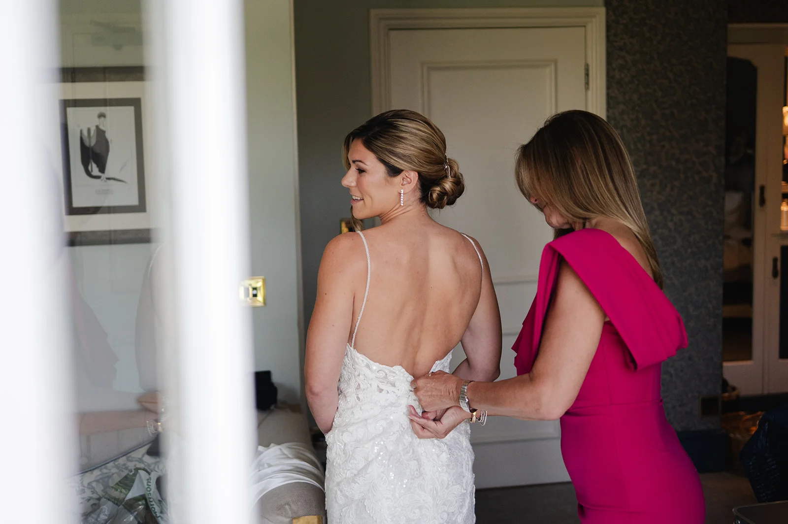 The mother of the bride gently zips up the back of the lace wedding dress as the smiling bride looks back over her shoulder.