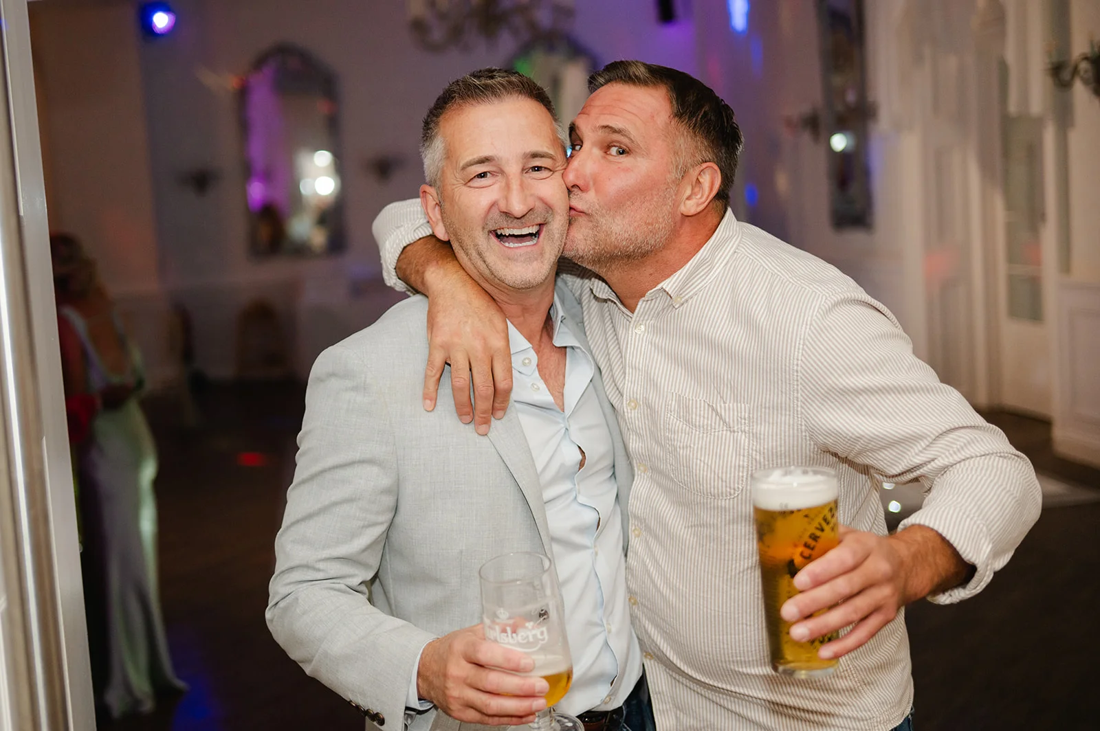 Two smiling male wedding guests embracing and sharing a joyful kiss while holding glasses of beer during the evening reception.