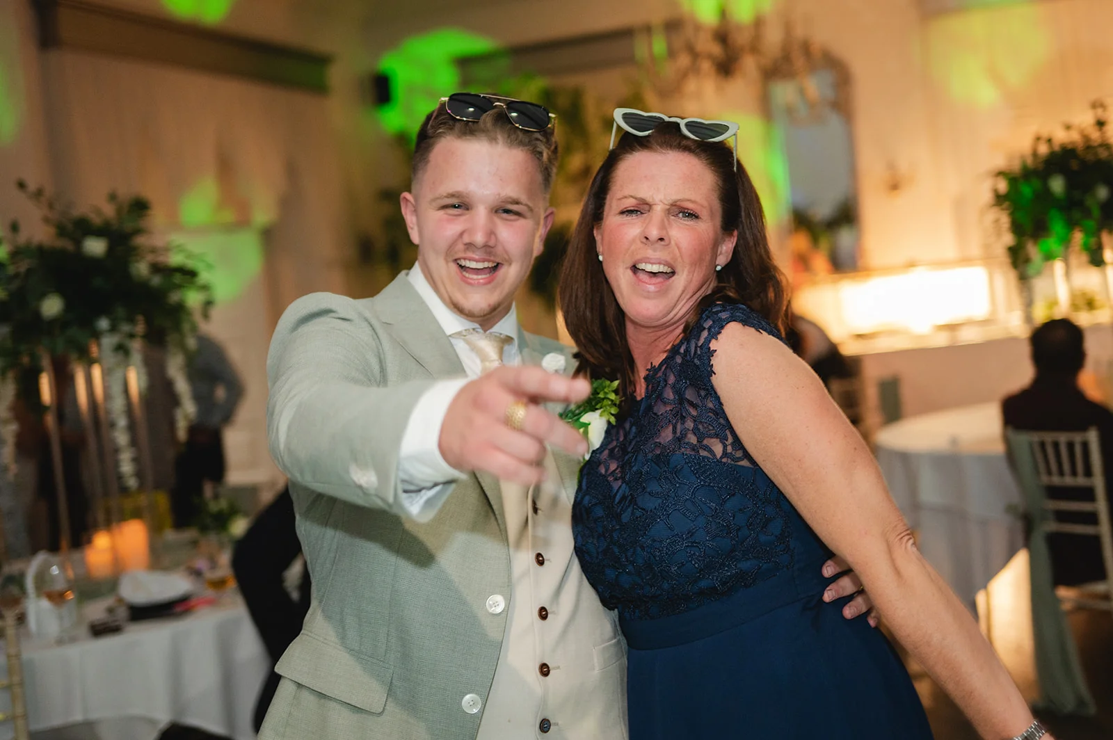 A groom in a sage green suit and a guest wearing sunglasses share a joyful, laughing moment on the dance floor during an evening wedding reception.