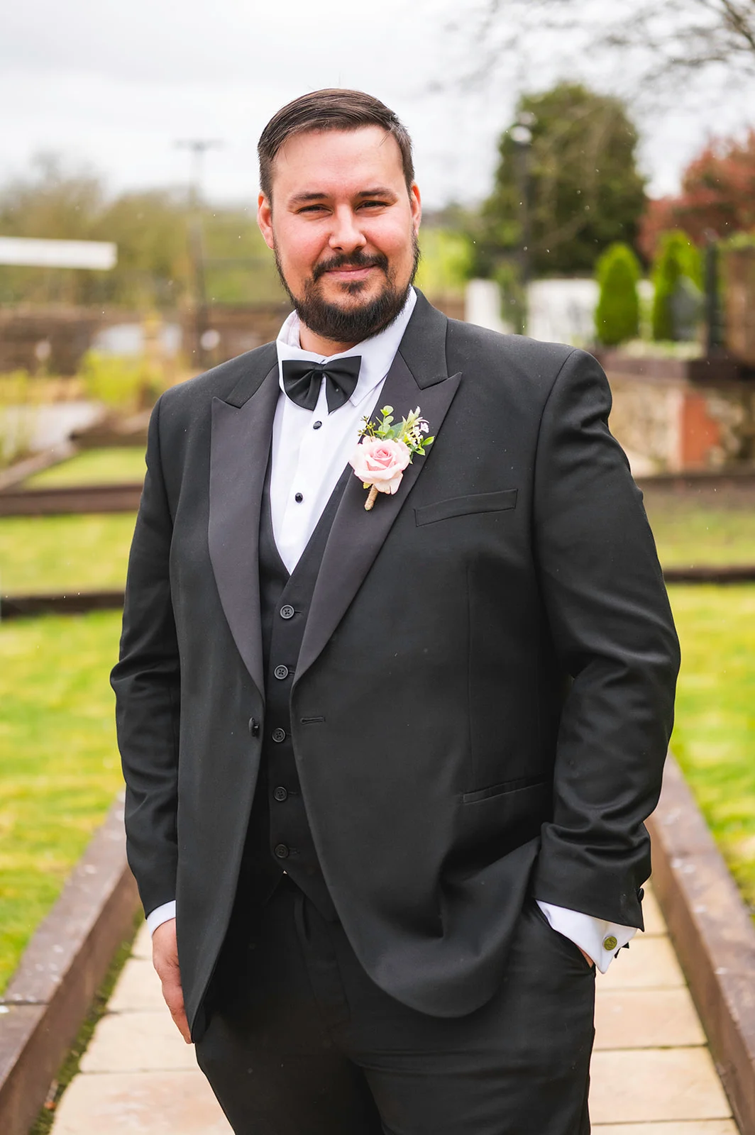 A smiling groom poses outdoors wearing a black tuxedo and matching waistcoat with a pink rose buttonhole pinned to his lapel.