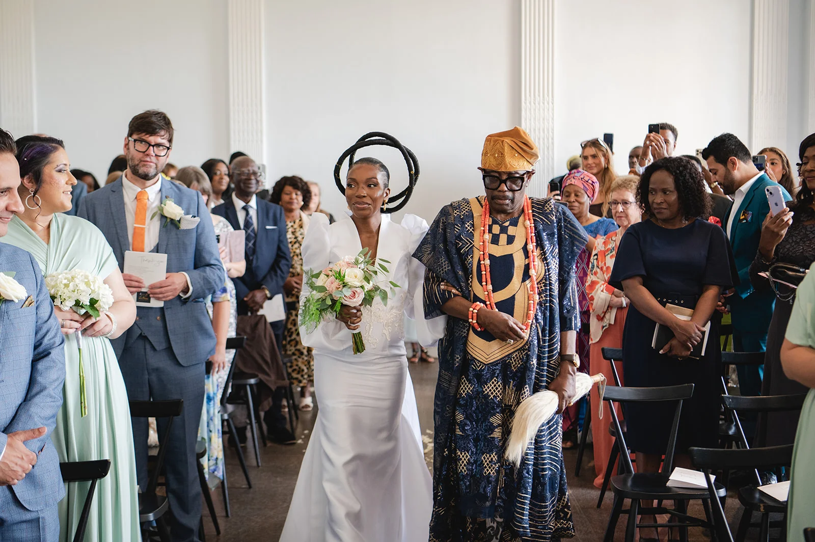 A bride in a white gown and striking halo headpiece walks down the aisle with her father, who wears traditional Nigerian attire and coral beads, while guests look on.