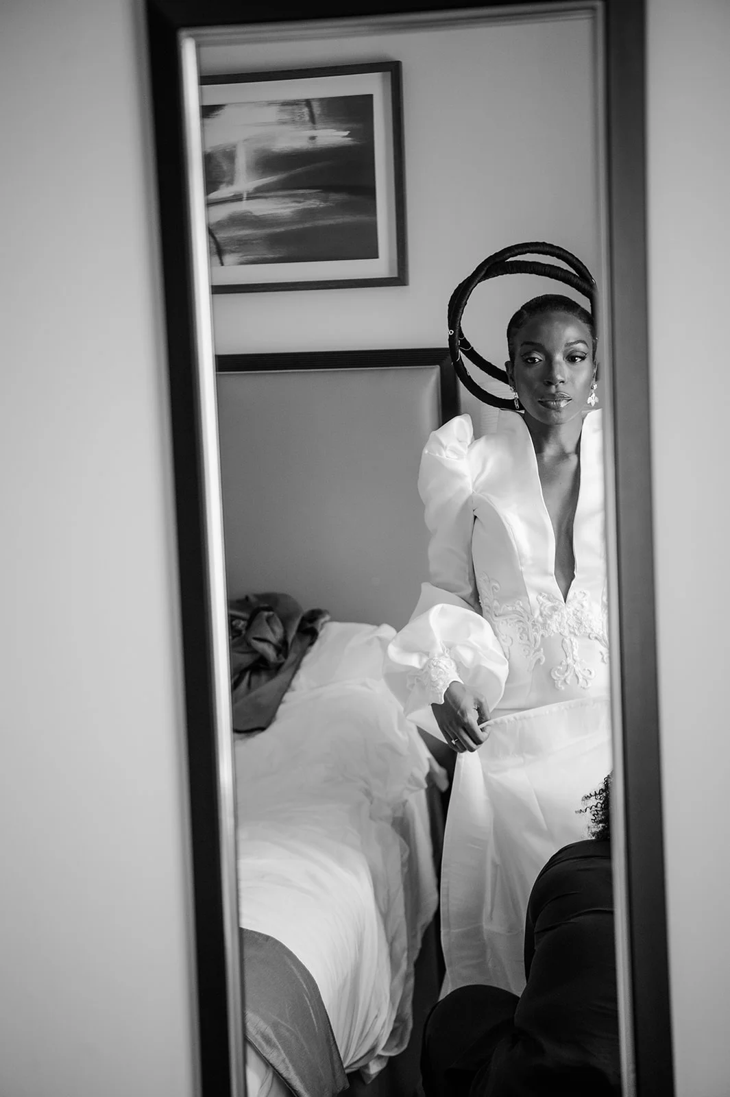 A black and white portrait of a bride looking into a mirror while getting ready, wearing an embroidered gown with statement headpiece.