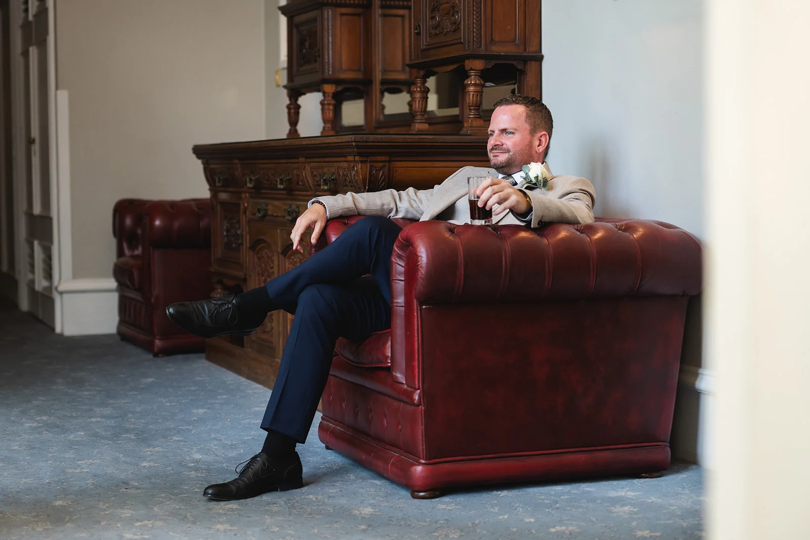 A relaxed groom with a buttonhole sits comfortably in a red leather armchair, holding a glass inside an elegant country house.