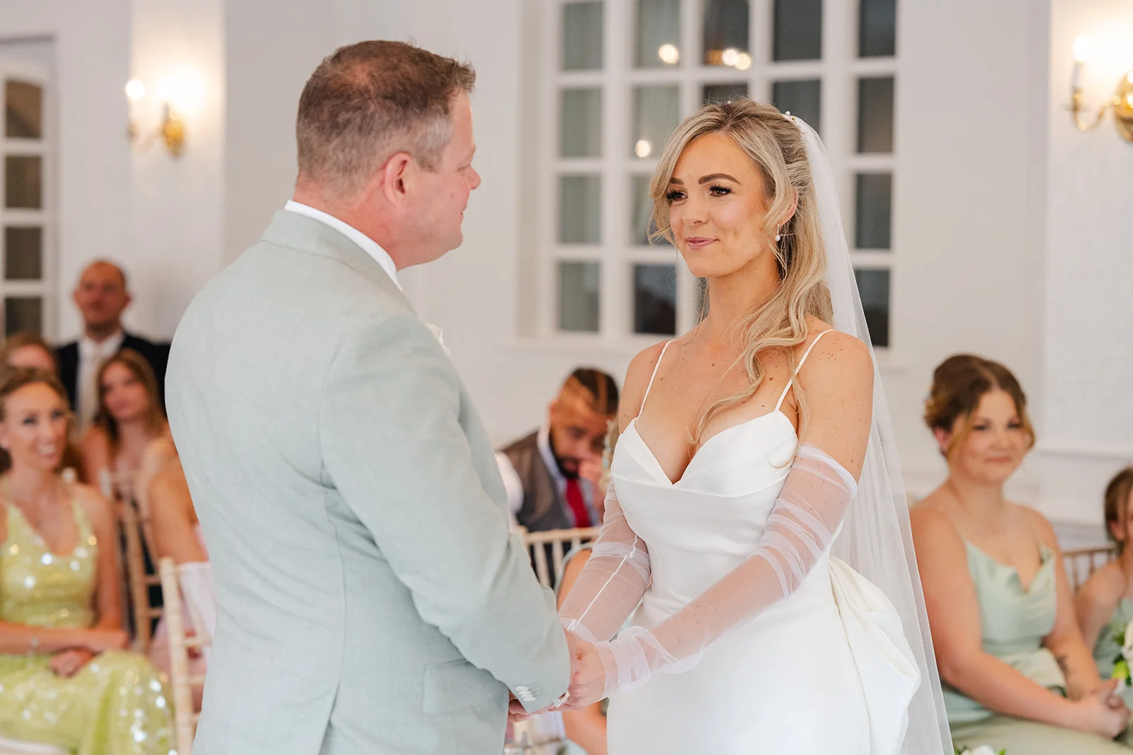 A newlywed couple holds hands during their civil wedding ceremony, with the smiling bride wearing a satin gown featuring a sweetheart neckline and sheer gloves.