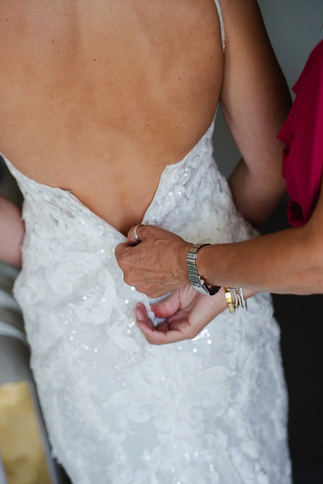 A close-up shot from behind showing a bride being helped into her intricate, beaded white lace wedding dress with spaghetti straps.