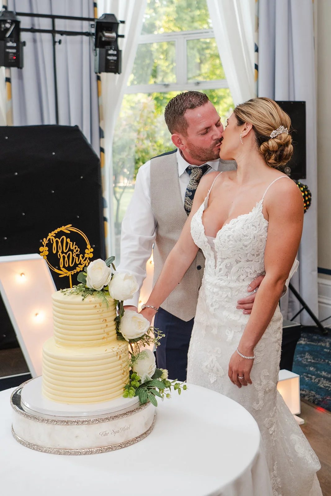 A bride and groom share a tender kiss while cutting their elegant two-tiered white wedding cake adorned with fresh roses and a gold topper.
