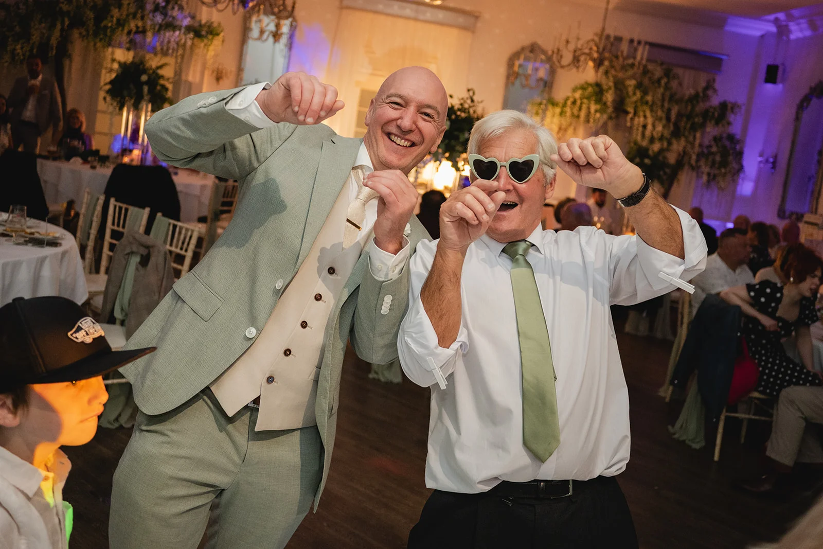Two smiling men in suits dance joyfully on a brightly lit evening dance floor, with one wearing novelty heart-shaped sunglasses.