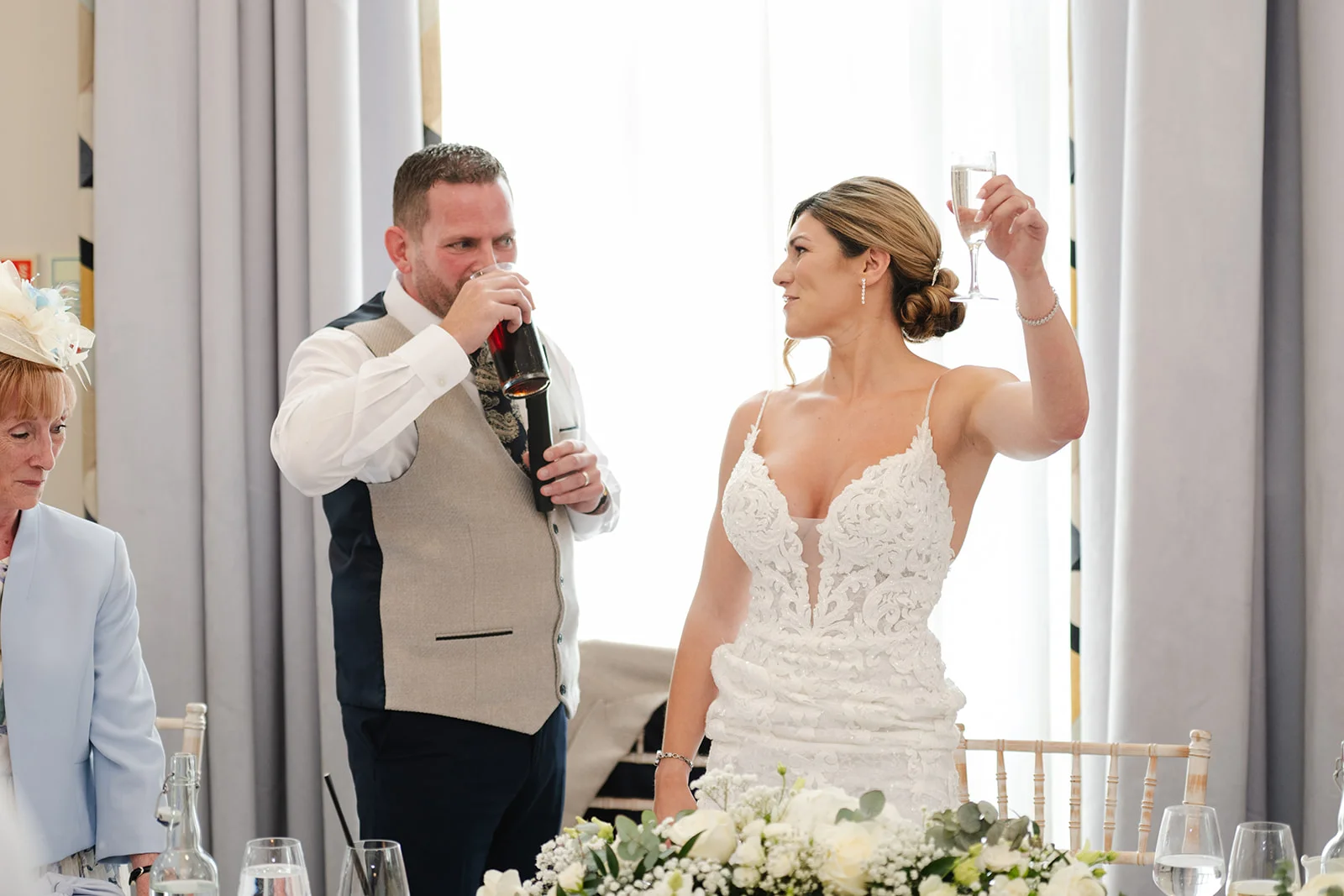 A bride in a plunging lace gown raises a toast with a glass of champagne while standing beside her groom at the wedding reception top table.