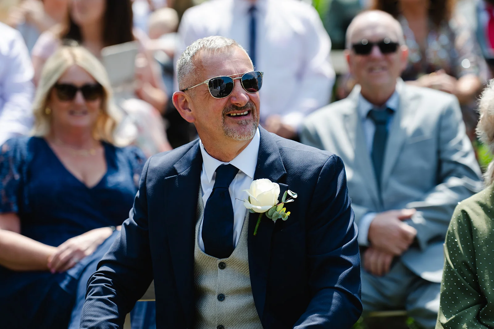 A smiling groom wearing a navy suit, tweed waistcoat, and sunglasses sits amongst wedding guests during the ceremony.