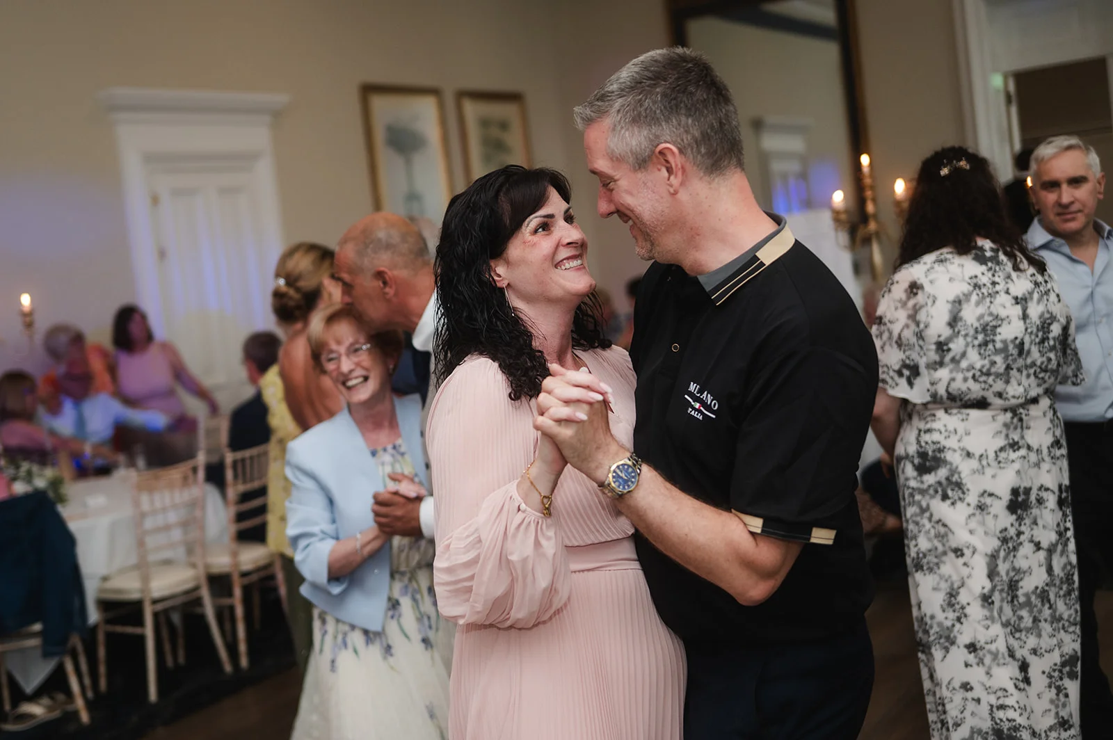 A mature couple smile lovingly at each other while dancing together at a lively evening wedding reception surrounded by family and guests.