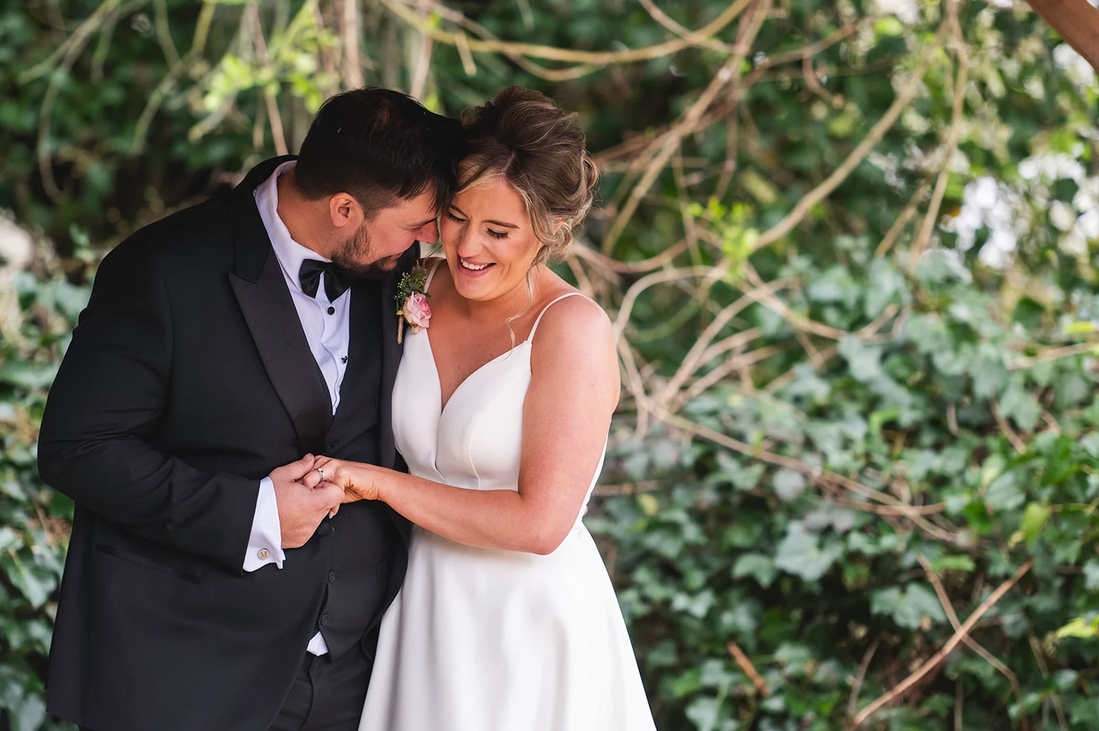A newlywed couple share a tender moment with the groom kissing the bride's forehead while they hold hands outdoors against a backdrop of greenery.