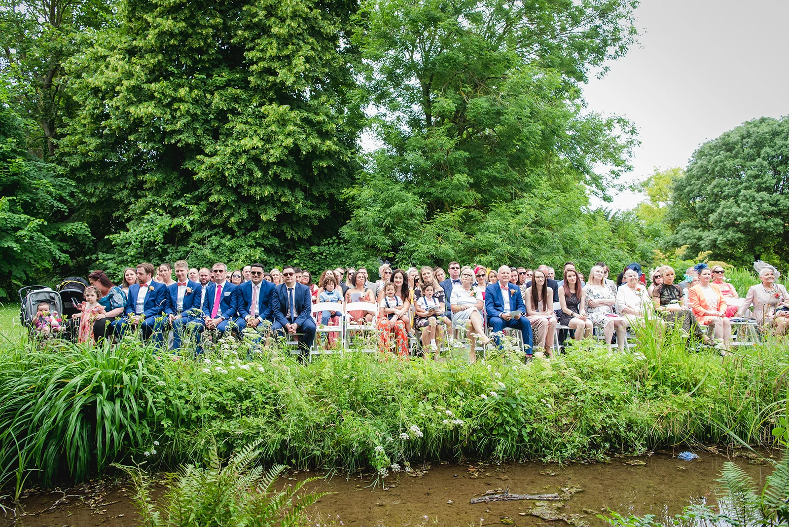 Wedding guests are seated in white chairs along a lush riverbank for an outdoor summer ceremony.