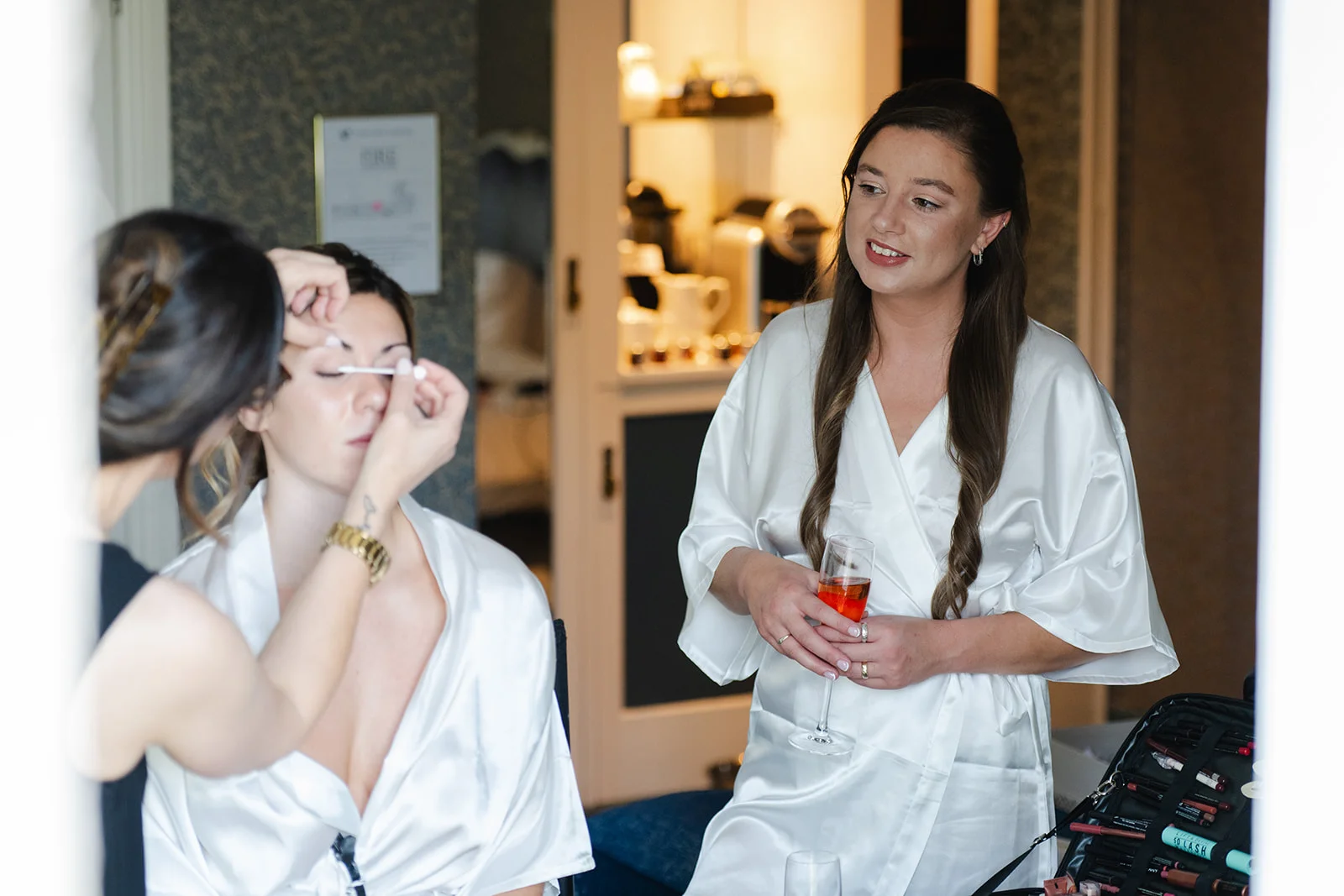 A smiling bridesmaid in a white satin robe holds a glass of champagne while another person gets her makeup applied in the background.