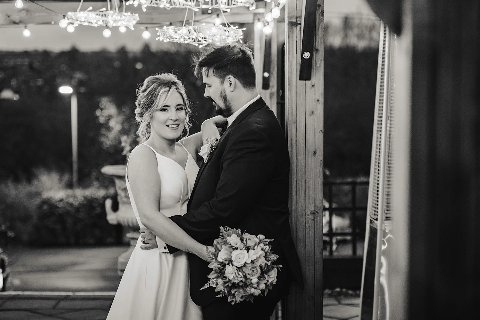 A newlywed couple shares a tender moment beneath an outdoor canopy illuminated by festoon lights at night.