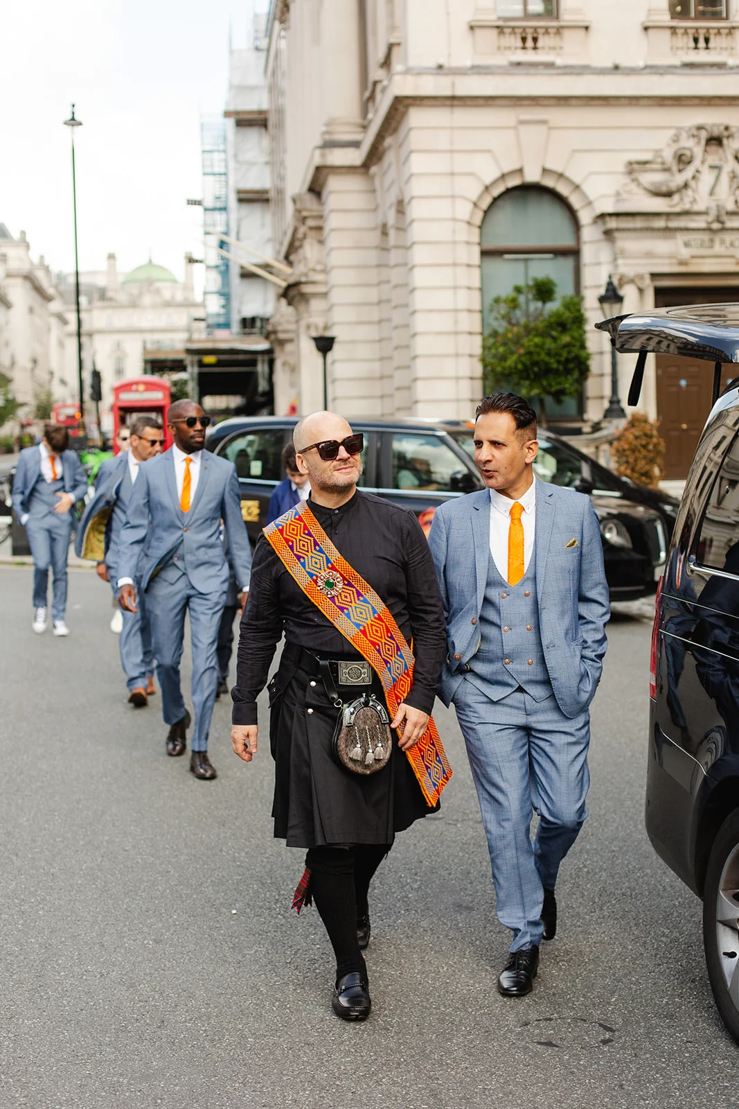 A groom wearing a black kilt and ornate colourful sash walks along a London street alongside groomsmen in light grey three-piece suits with orange ties.