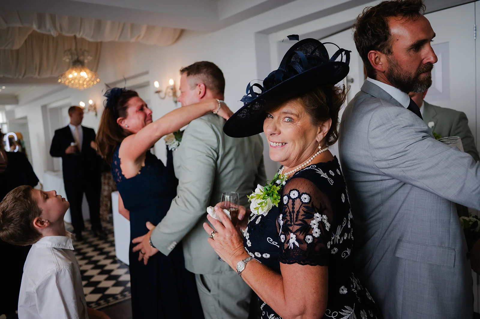 A smiling guest wearing a navy fascinator and floral dress looks back towards the camera while wedding guests mingle in the background.