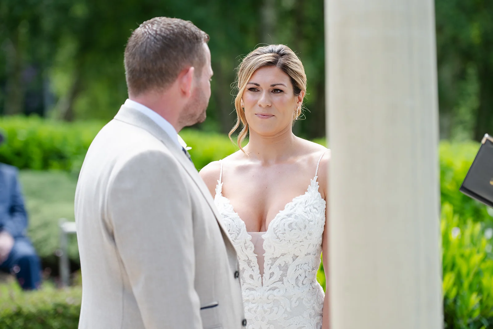 A groom in a light suit looks lovingly at his bride wearing an intricately patterned lace wedding dress with a plunging neckline during an outdoor ceremony.