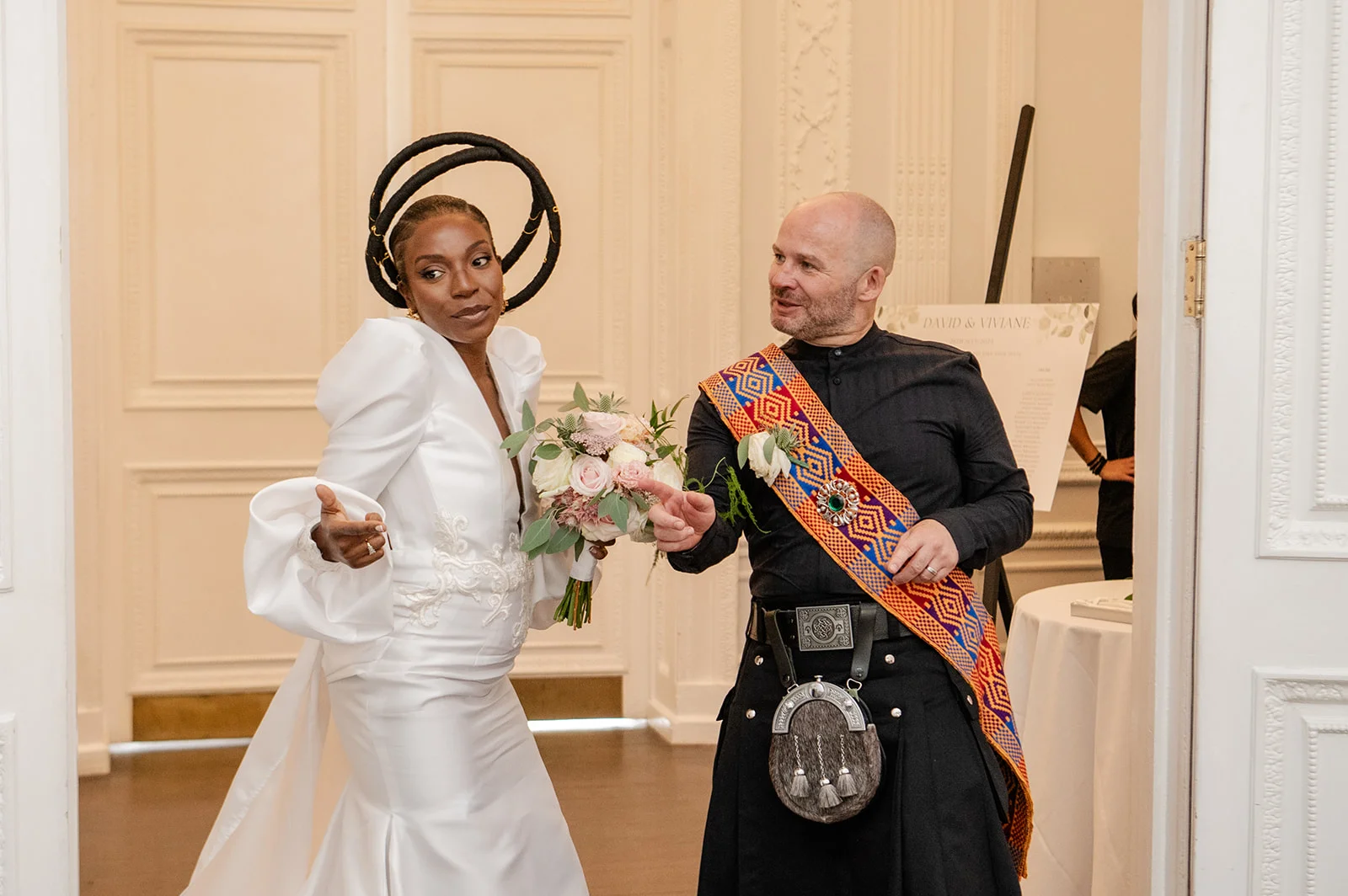 A newlywed couple laughing together in an elegant room, with the bride wearing a white gown featuring dramatic puff sleeves and a sculptural halo headpiece, while the groom wears a traditional black kilt with a patterned sash.