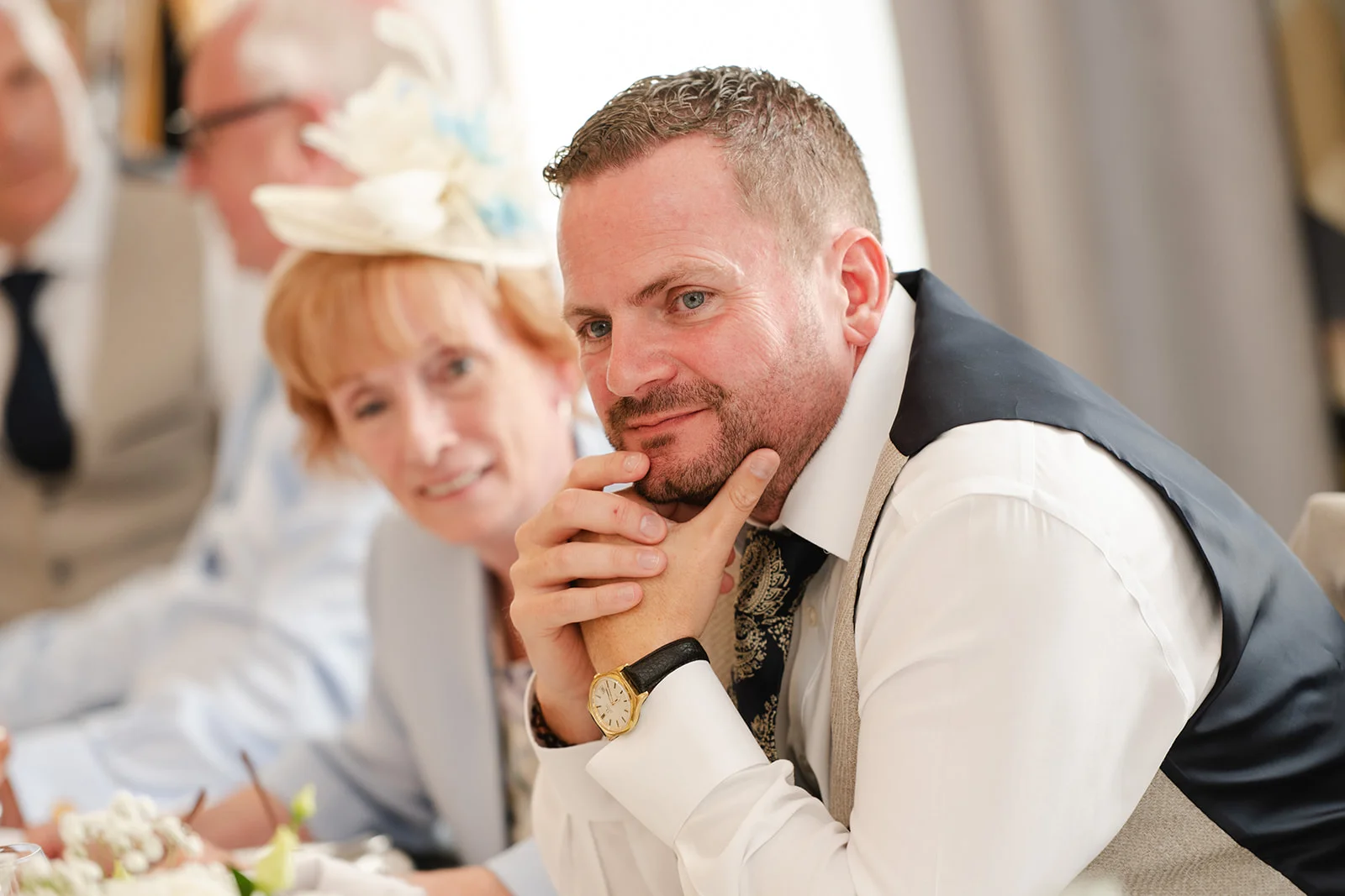 A groom sitting at the reception table with his chin on his clasped hands, attentively watching the wedding speeches.