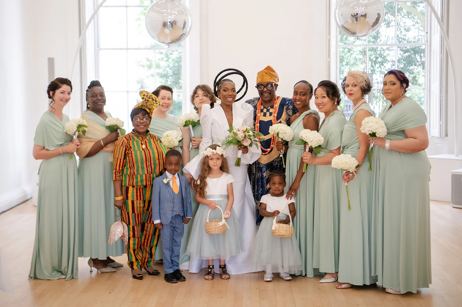 The newly married couple pose with their large bridal party wearing sage green dresses and traditional attire in a bright, modern venue.