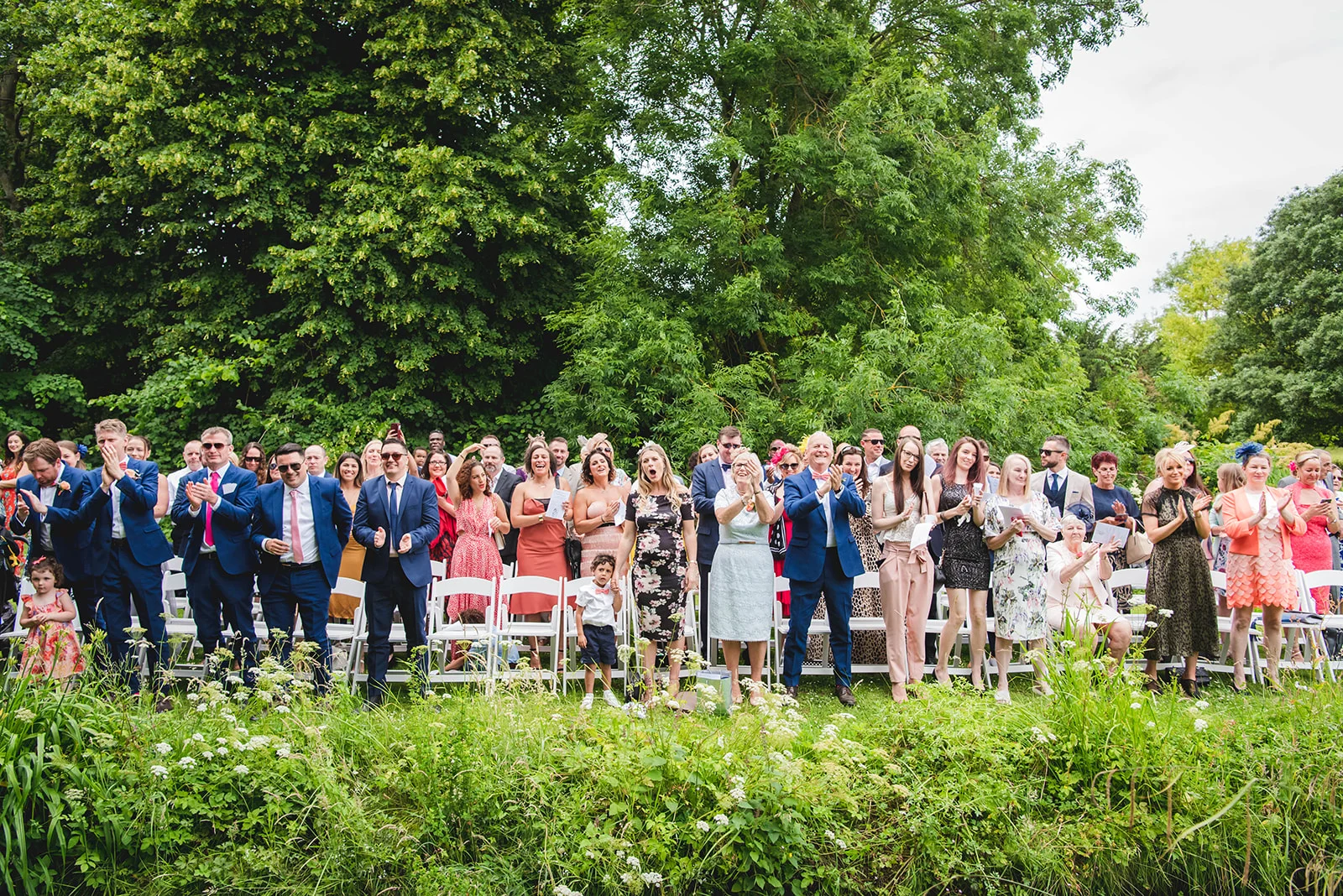 A joyful crowd of wedding guests sitting and standing outdoors in a lush garden setting, applauding and cheering with smiles.
