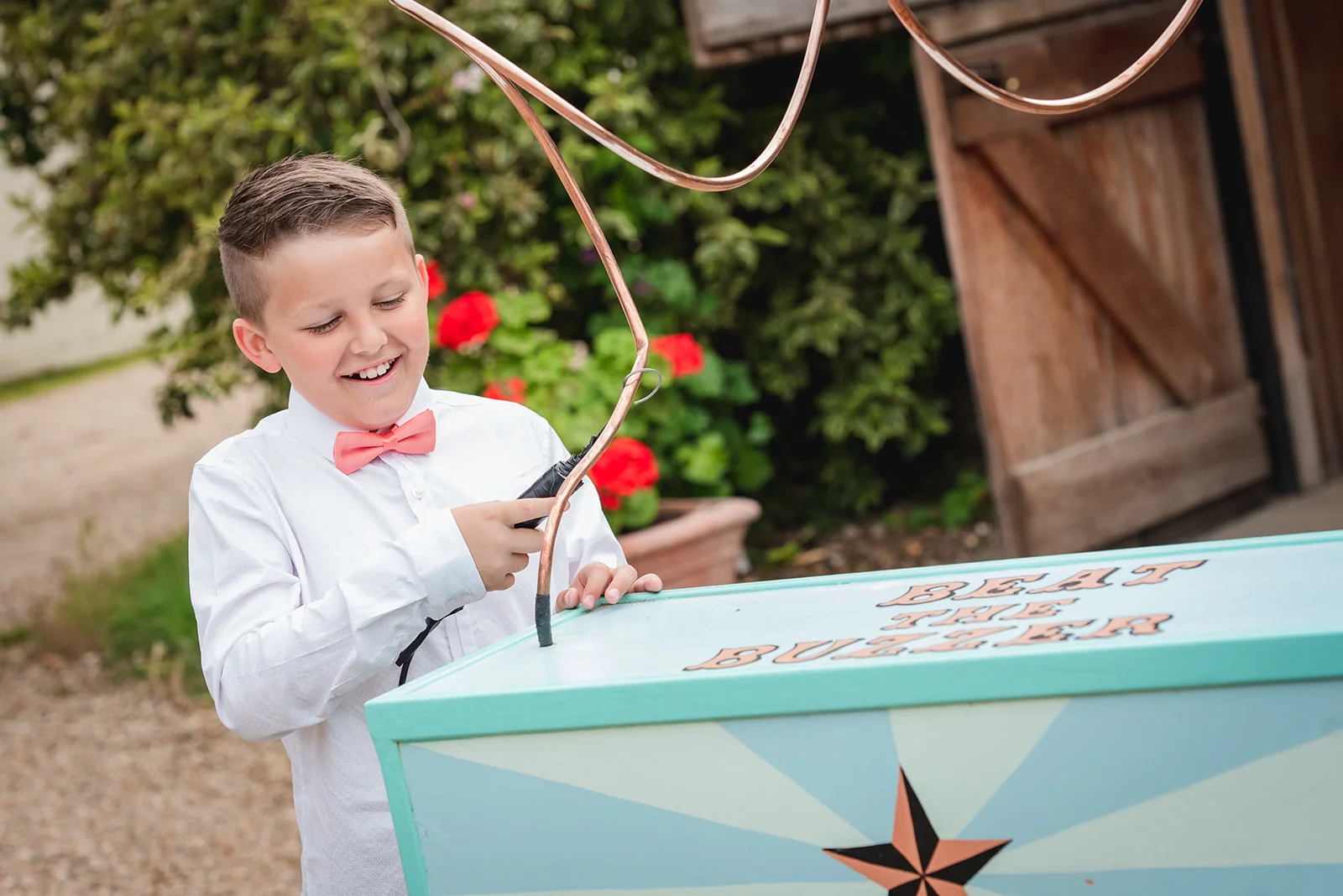 A smiling page boy in a white shirt and pink bow tie concentrates on playing a beat the buzzer lawn game outdoors near a rustic barn.