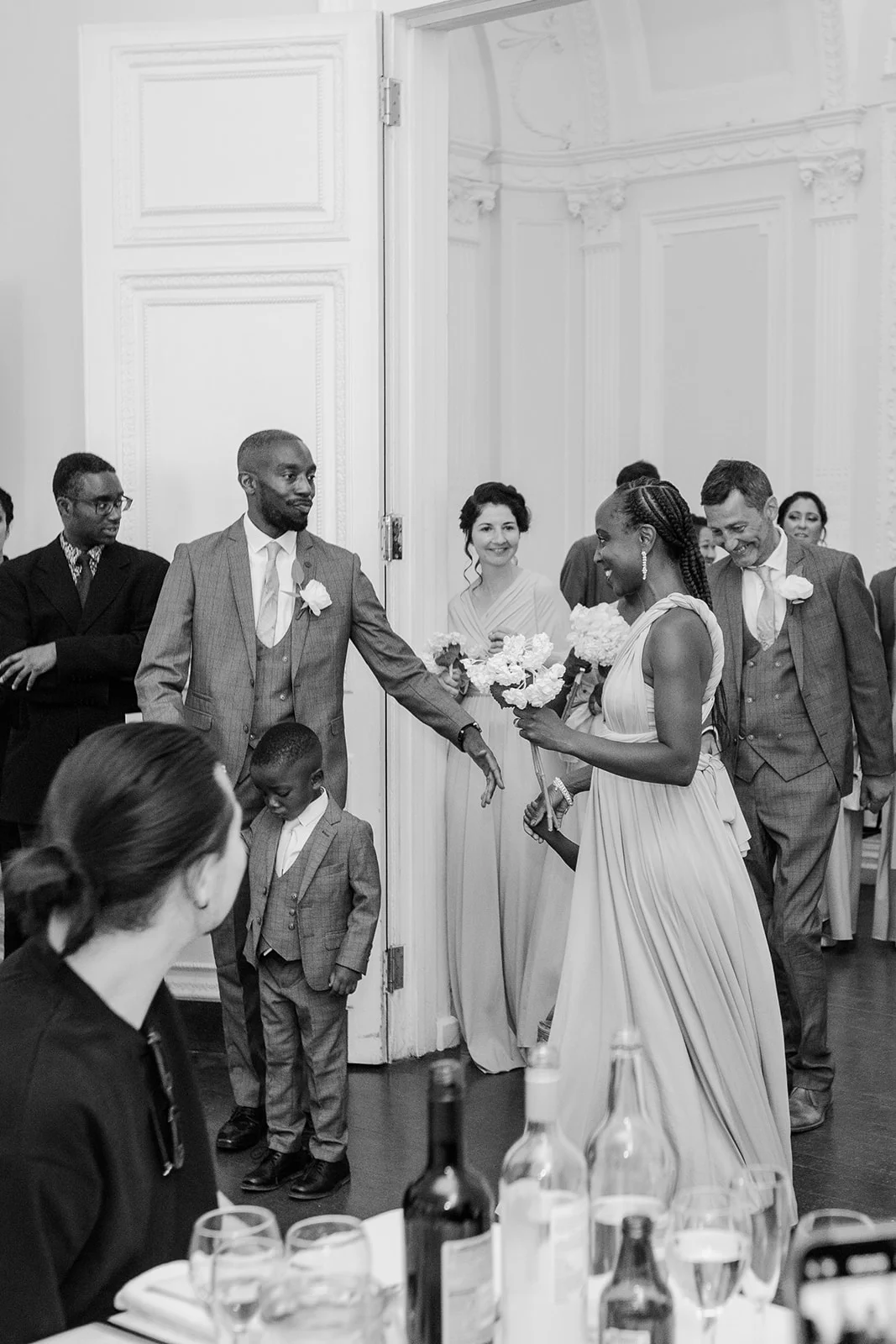 A candid black and white photograph captures wedding guests and a page boy entering a bright reception room through open double doors, smiling and holding small bouquets.