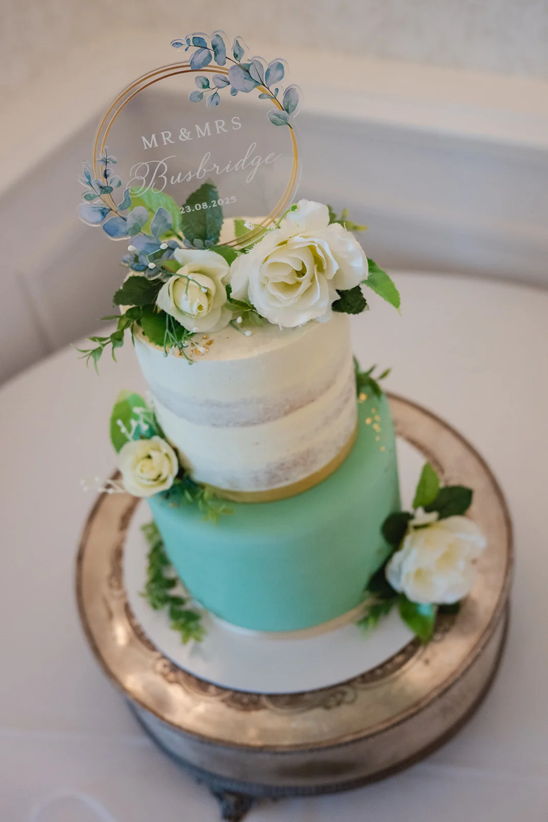 A two-tier wedding cake features a mint green bottom tier and a semi-naked top tier, adorned with white roses and a personalised acrylic ring topper.