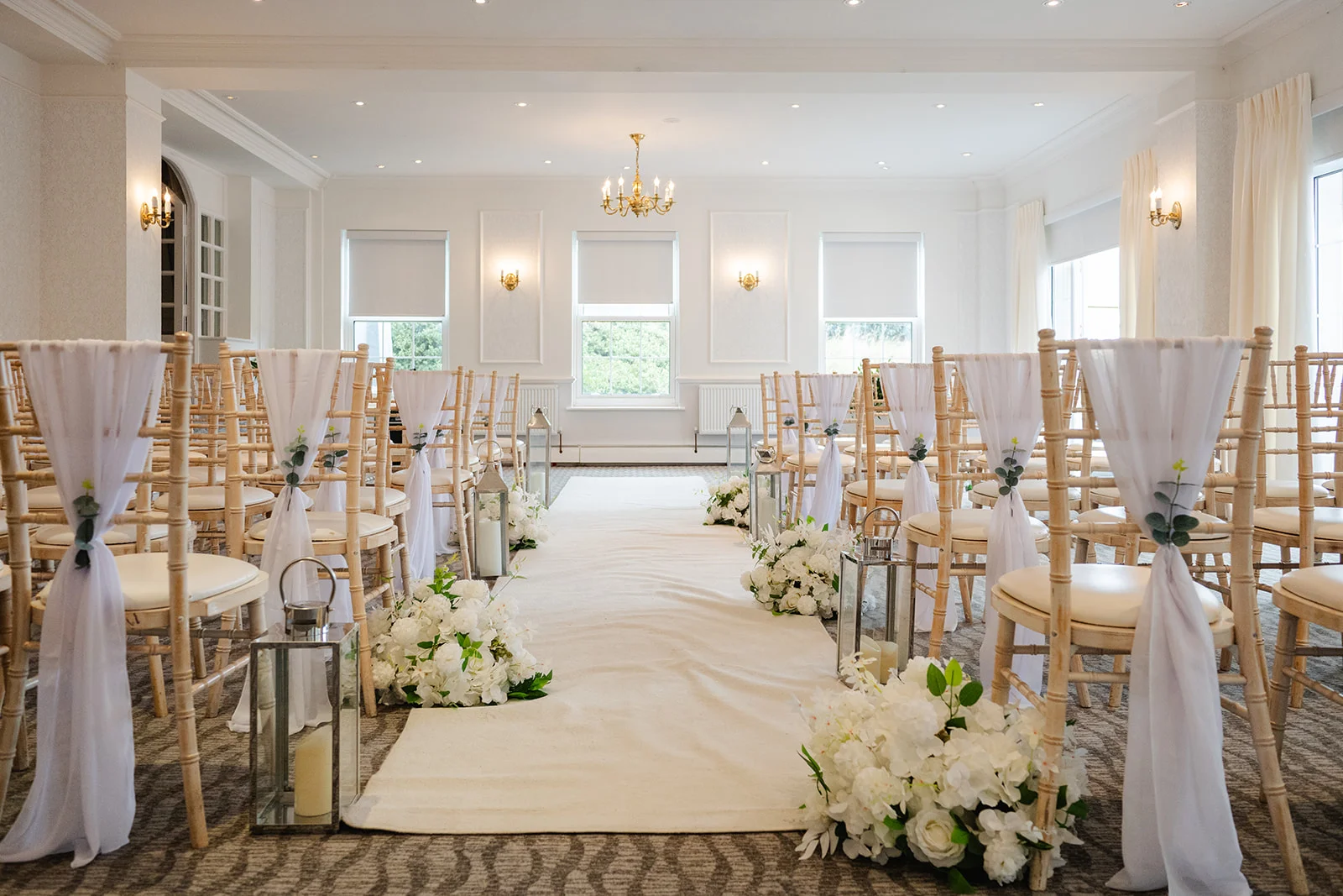 An elegant hotel wedding ceremony room features light wood chairs draped with white fabric, candle lanterns, and lush white floral arrangements lining the aisle.